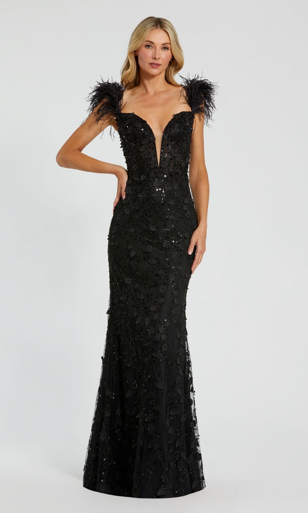 A woman showcases the Long Formal Dress 20889 by Mac Duggal, featuring a fitted, floor-length black silhouette with a sweetheart v-neckline, sequin details, and bold feather straps against a plain background.